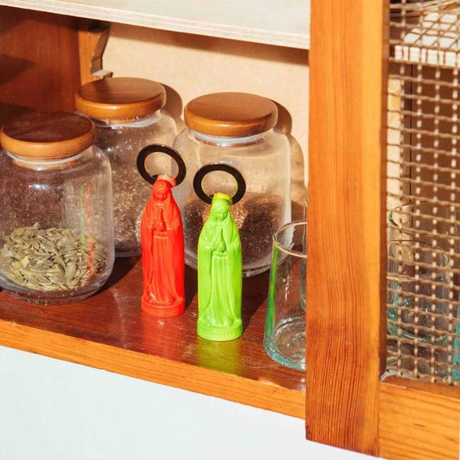 Otwieracz do butelek zielony w kształcie figurki stojący na półce obok czerwonego otwieracza i szklanych pojemników