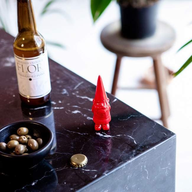 Czerwony otwieracz do butelek krasnal na czarnym marmurowym blacie obok butelki piwa i miski z oliwkami