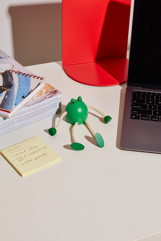 drewniana figurka żaba zielona z ruchomymi kończynami na biurku obok laptopa i notatek