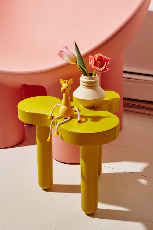 Drewniana figurka żyrafa od Areaware siedząca na żółtym stoliku z wazonem i kwiatami