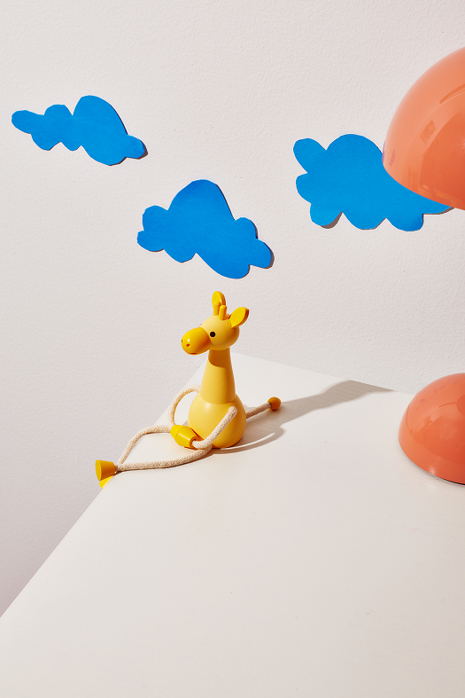Drewniana figurka żyrafa w żółtym kolorze z sznurkowymi nogami i niebieskimi chmurkami w tle