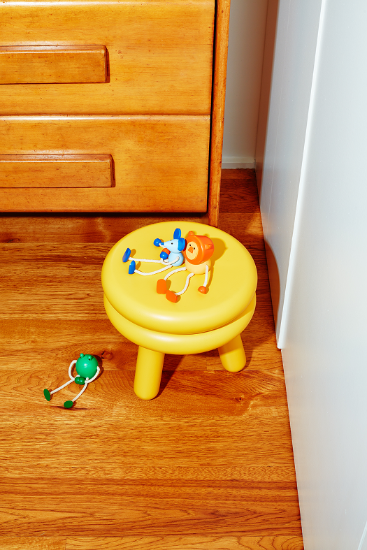 Drewniana figurka żaba leżąca na drewnianej podłodze obok żółtego stołka z zabawkami na górze
