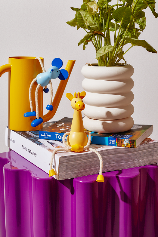 Drewniana figurka żyrafa stojąca na książkach obok żółtego dzbanka i białej doniczki z zieloną rośliną