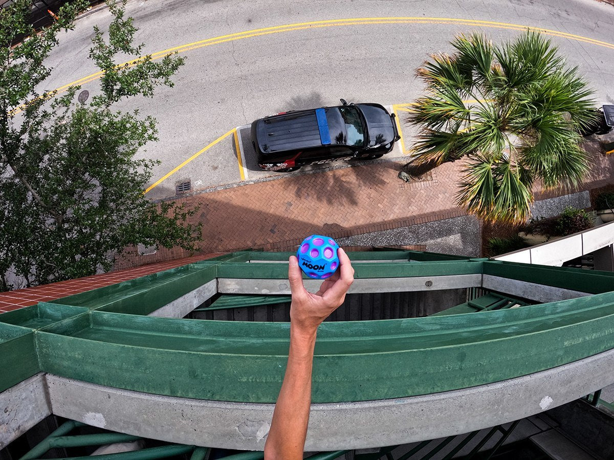 Ręka trzymająca piłeczkę Moon Ball Waboba nad barierką na tle ulicy i zaparkowanego samochodu