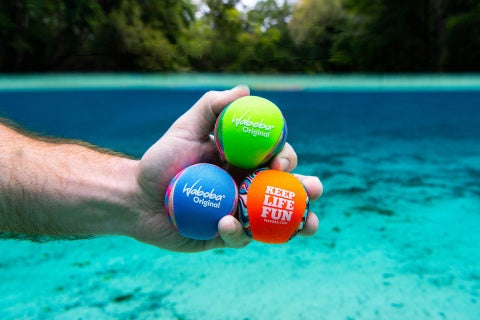 Trzy kolorowe piłeczki Waboba Original trzymane w dłoni nad wodą do zabawy na świeżym powietrzu