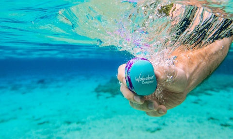 Ręka trzyma pod wodą niebiesko-fioletową piłeczkę Waboba Original do zabawy w wodzie