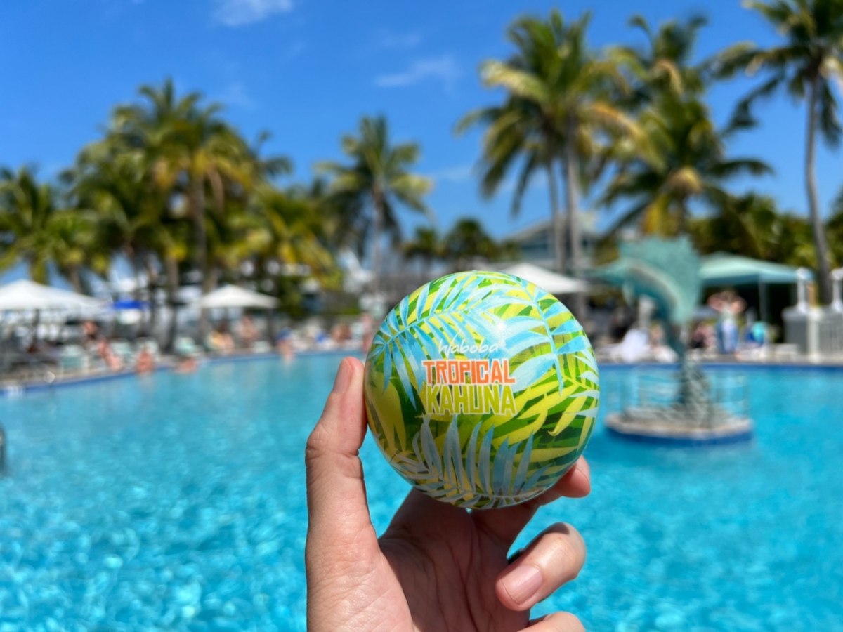 Piłka na wodę Waboba Tropical Kahuna w tropiczny wzór trzymana nad basenem z palmami w tle