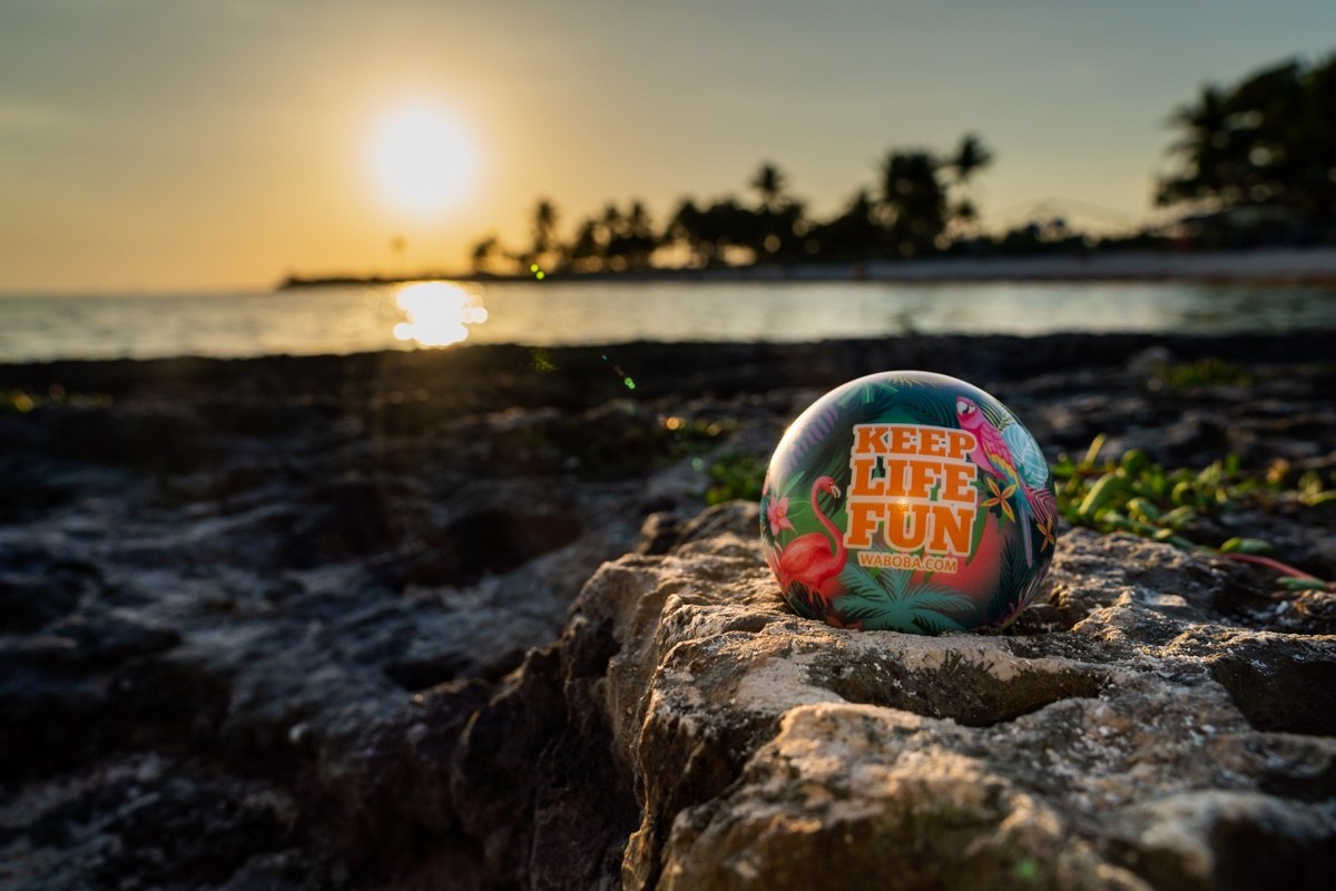 Piłka na wodę Waboba Tropical Kahuna z kolorowym wzorem flamingów na kamienistej plaży o zachodzie słońca