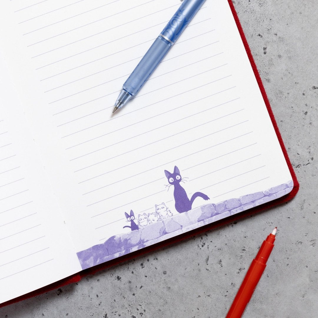Otwarty pluszowy notatnik Jiji z ilustracją fioletowych kotów i długopisy na szarym tle