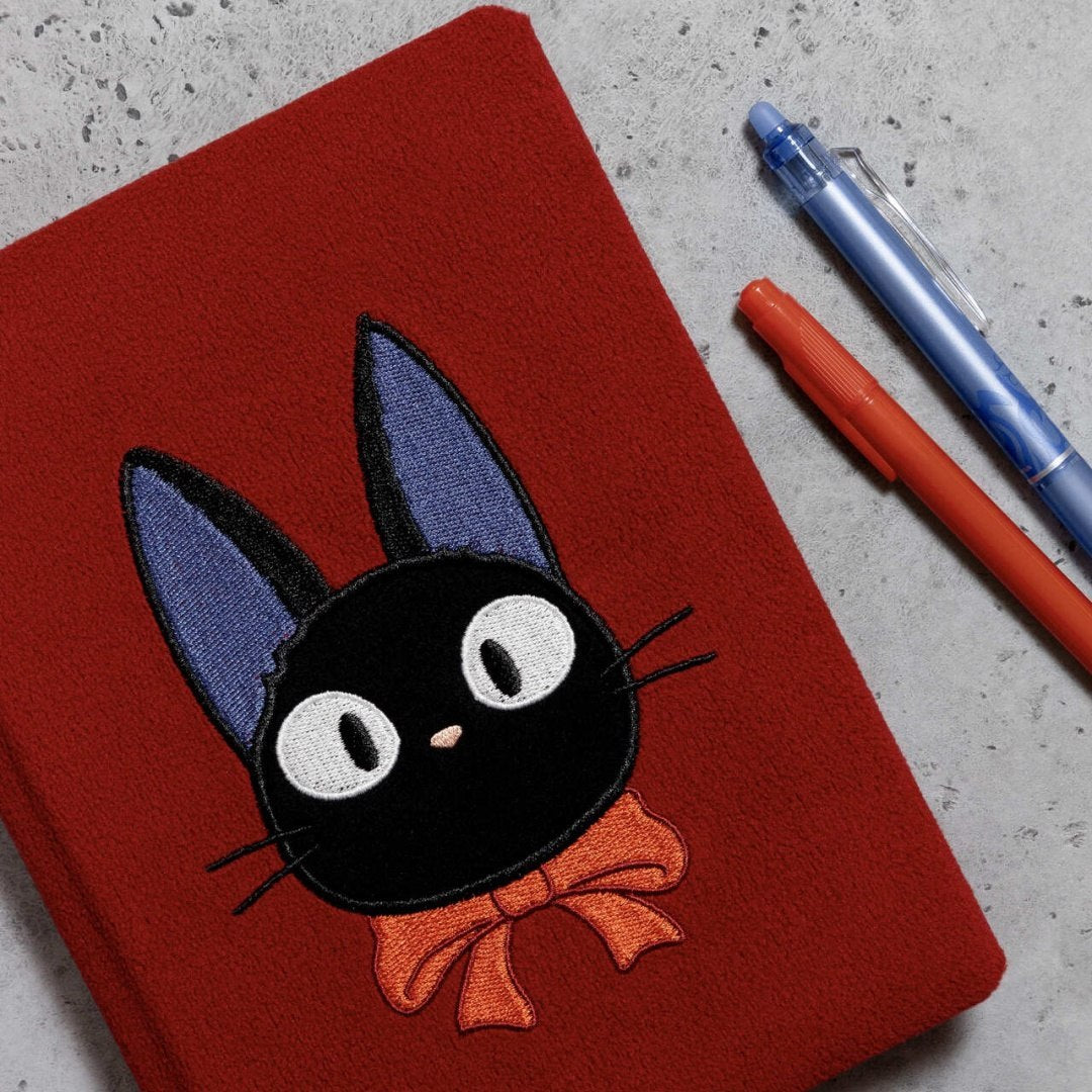 Czerwony pluszowy notatnik Jiji z haftowaną czarną postacią kota i pomarańczową kokardą obok długopisów