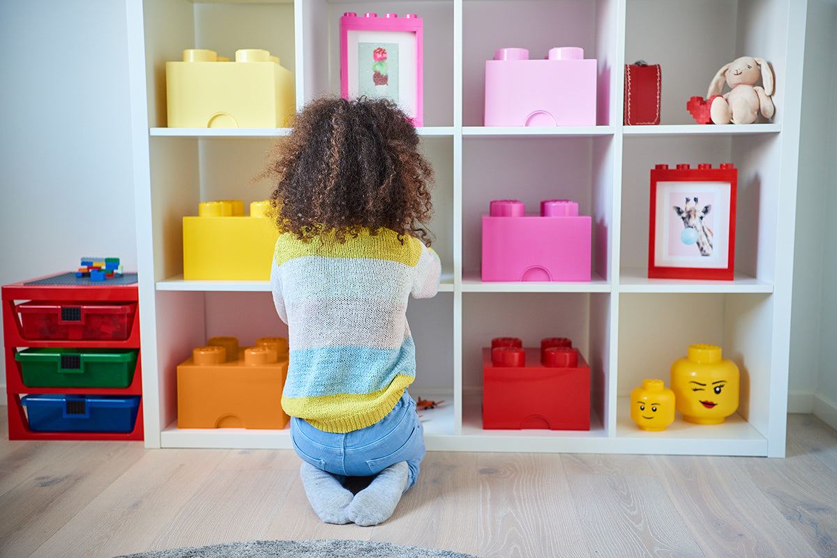 Pojemnik LEGO różowy w kształcie klocka na półce w pokoju dziecięcym obok innych kolorowych pojemników LEGO