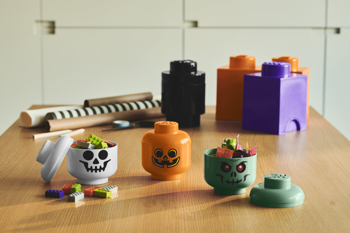 Pojemnik LEGO głowa zombie w zielonym kolorze z klockami na drewnianym stole obok innych pojemników LEGO