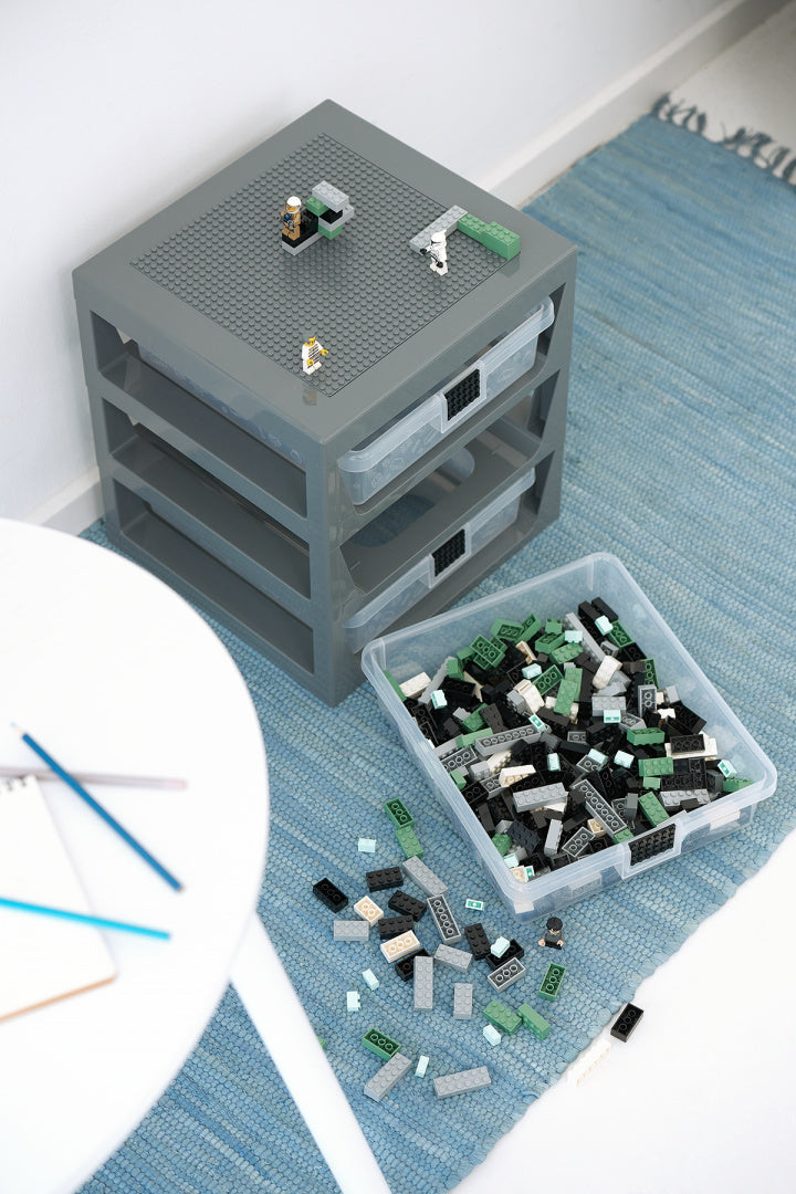 Szary regał LEGO z szufladami na klocki LEGO ustawiony na niebieskim dywanie obok stołu