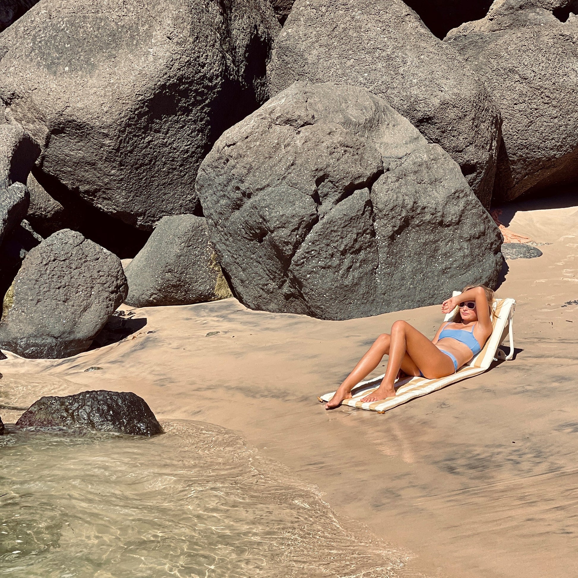 Kobieta opalająca się na rozkładanym leżaku plażowym Sunny Life na piaszczystej plaży przy skałach