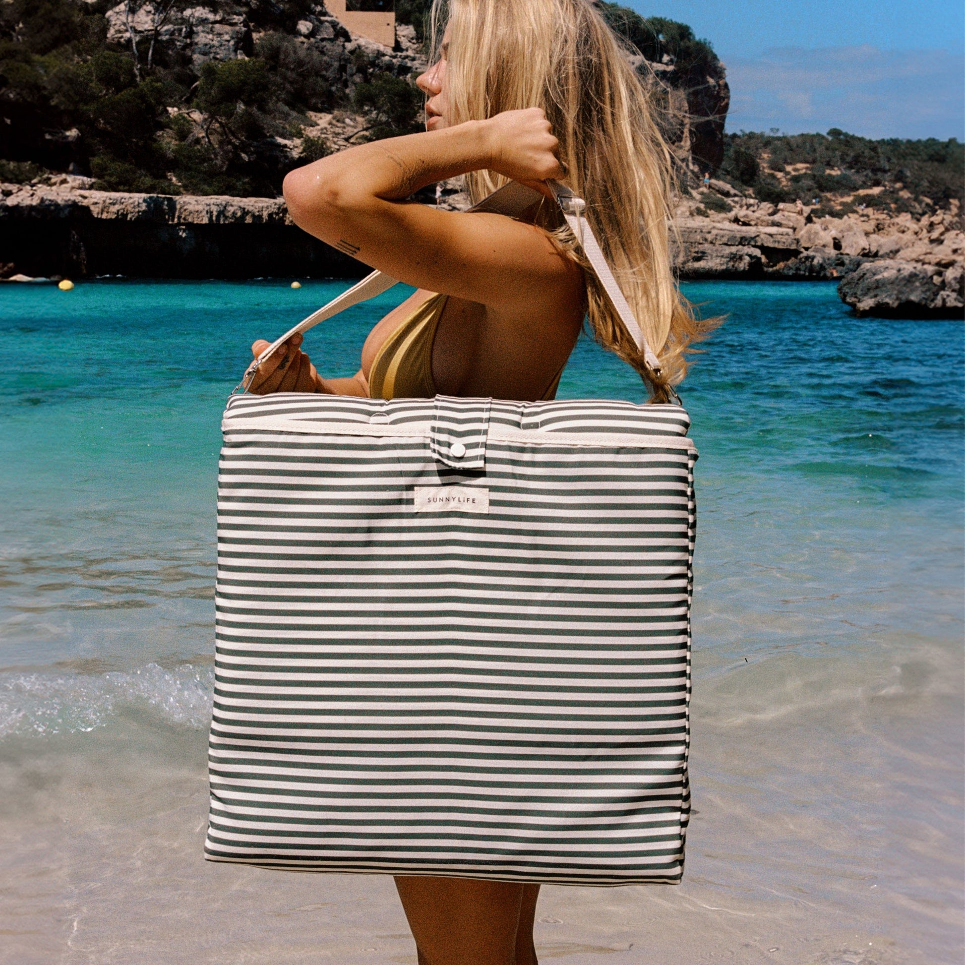 Kobieta niosąca torbę w paski z rozkładanym leżakiem plażowym Sunny Life na tle plaży i morza
