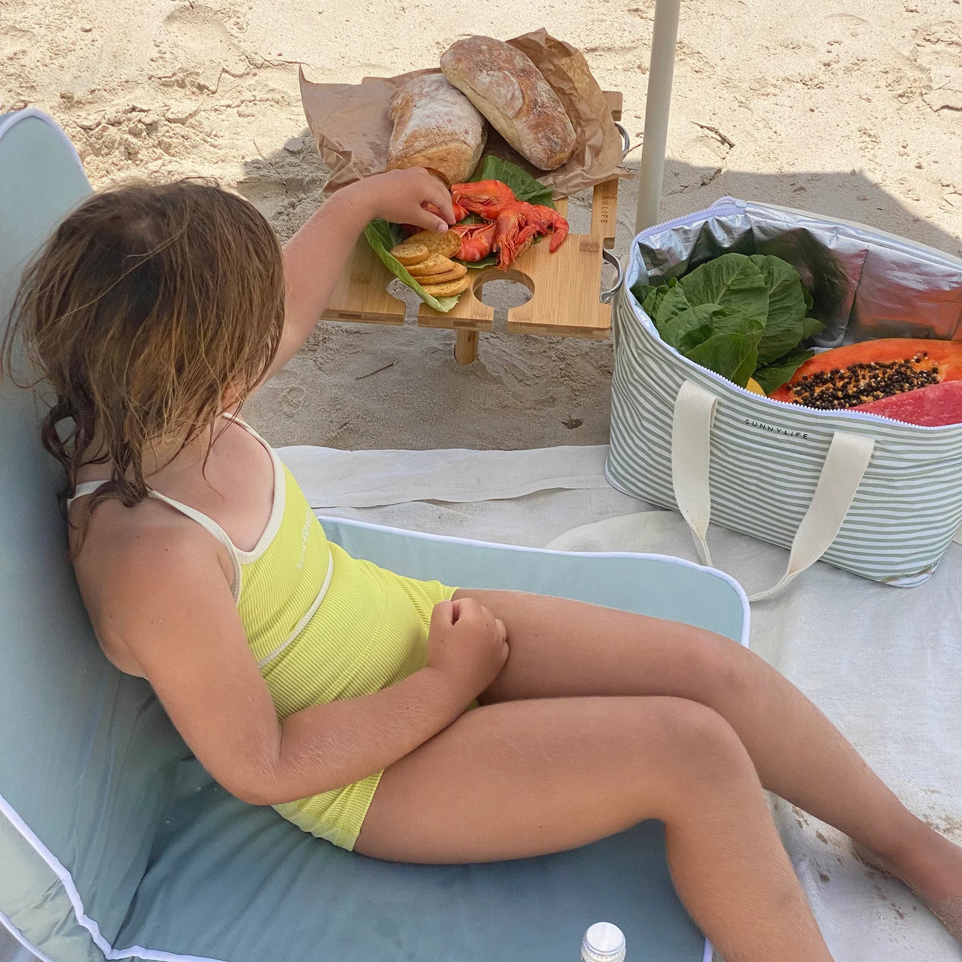 Dziecko na plaży siedzi na przenośnym stole piknikowym marki Sunny Life z przekąskami i owocami