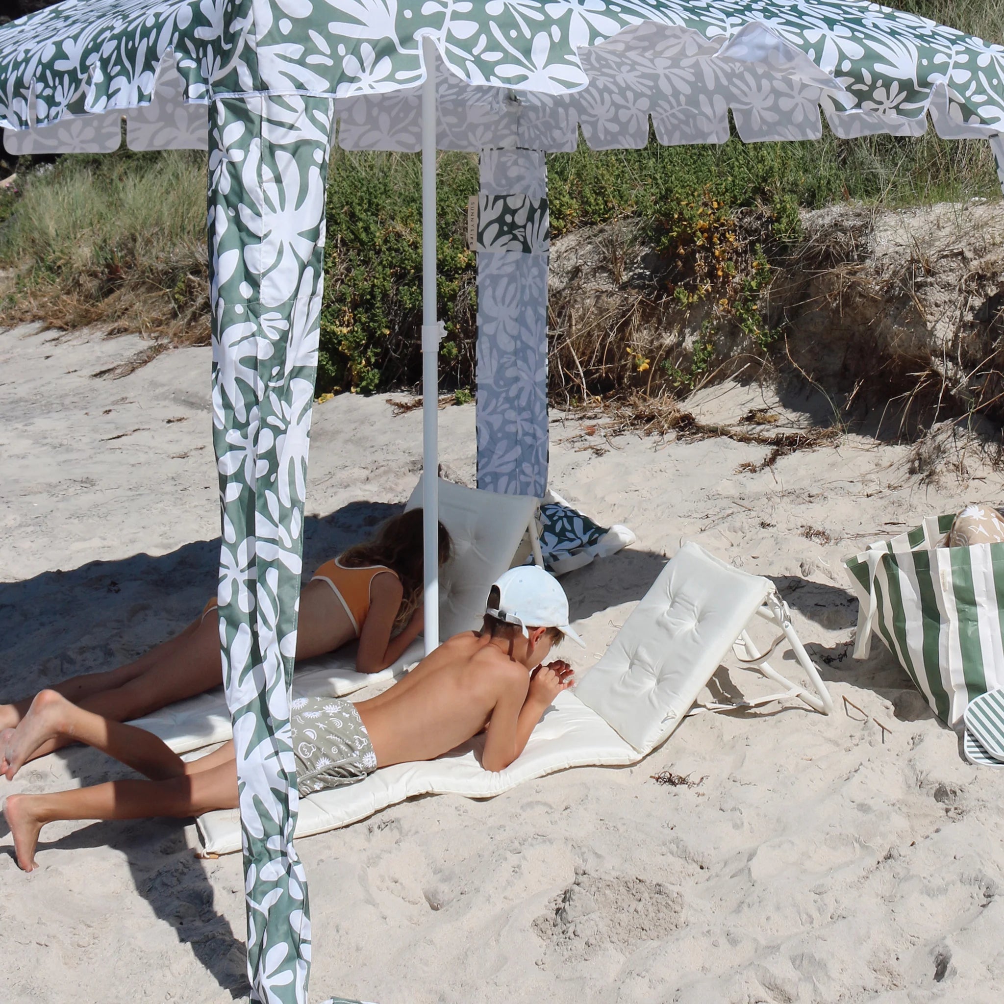 Dziecko leżące na białym rozkładanym leżaku plażowym pod zielono-białym parasolem na plaży