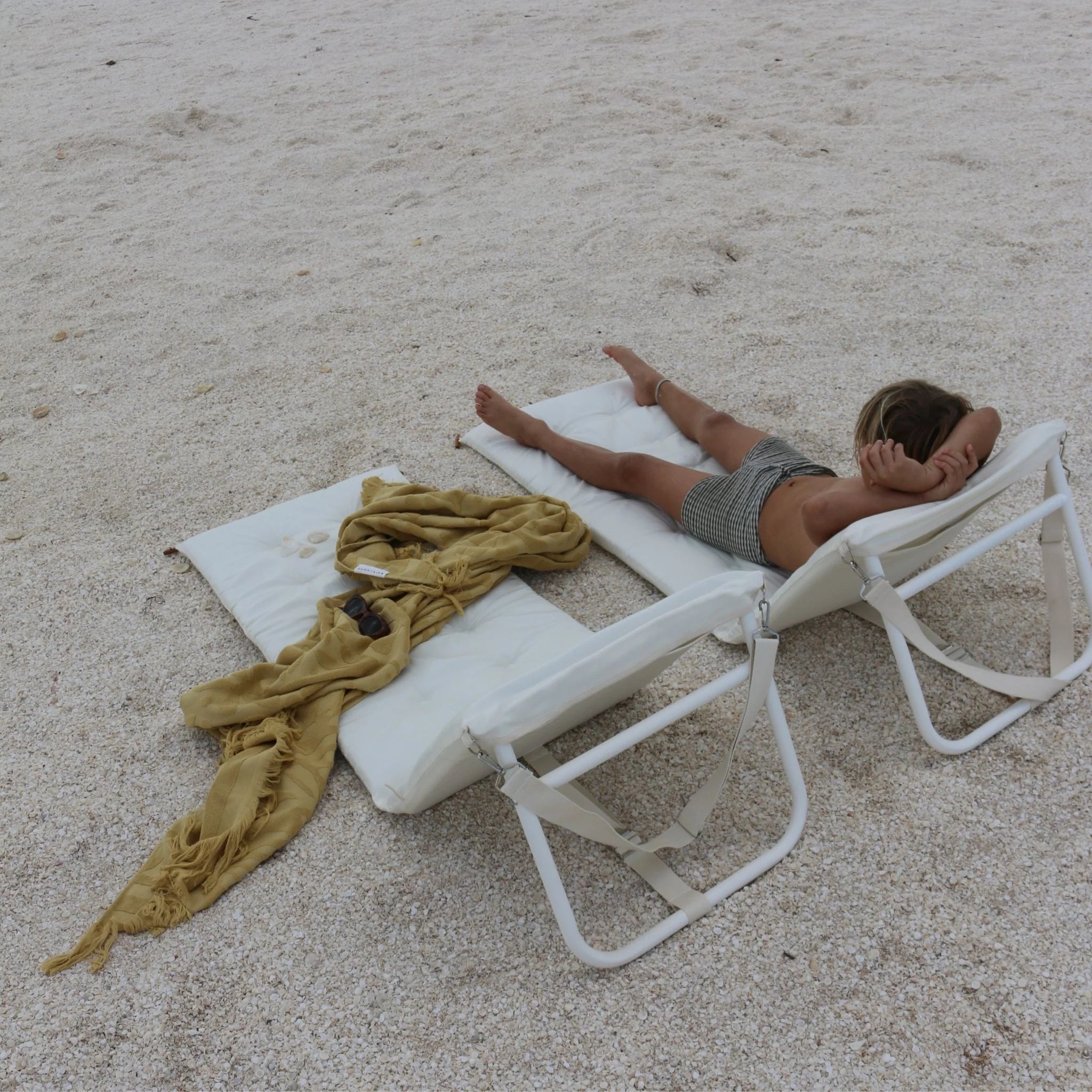 Biały rozkładany leżak plażowy Sunny Life z dzieckiem leżącym na piasku i żółtym ręcznikiem