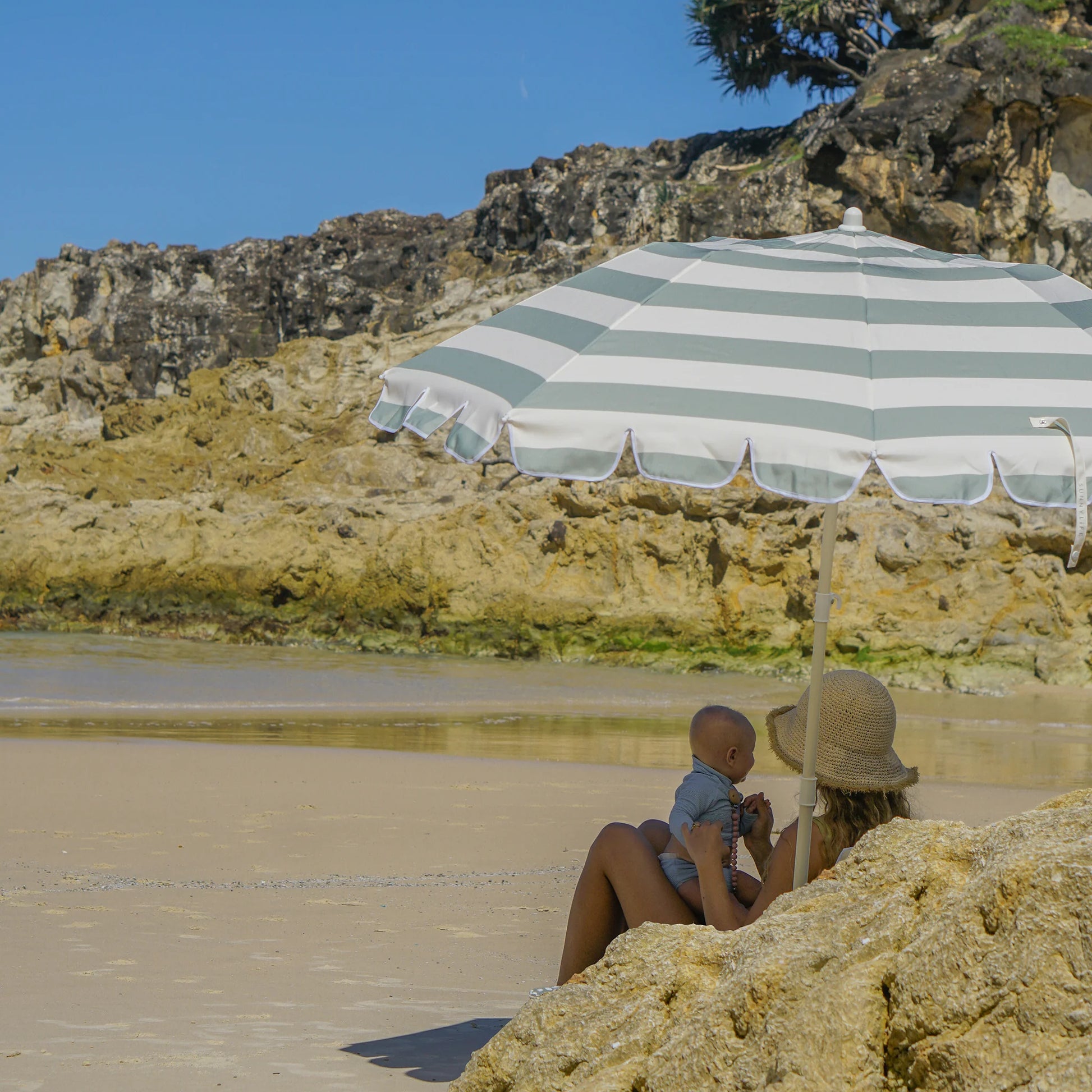 Kobieta z dzieckiem pod szarym paskowanym kompaktowym parasolem plażowym marki LOQI na piaszczystej plaży