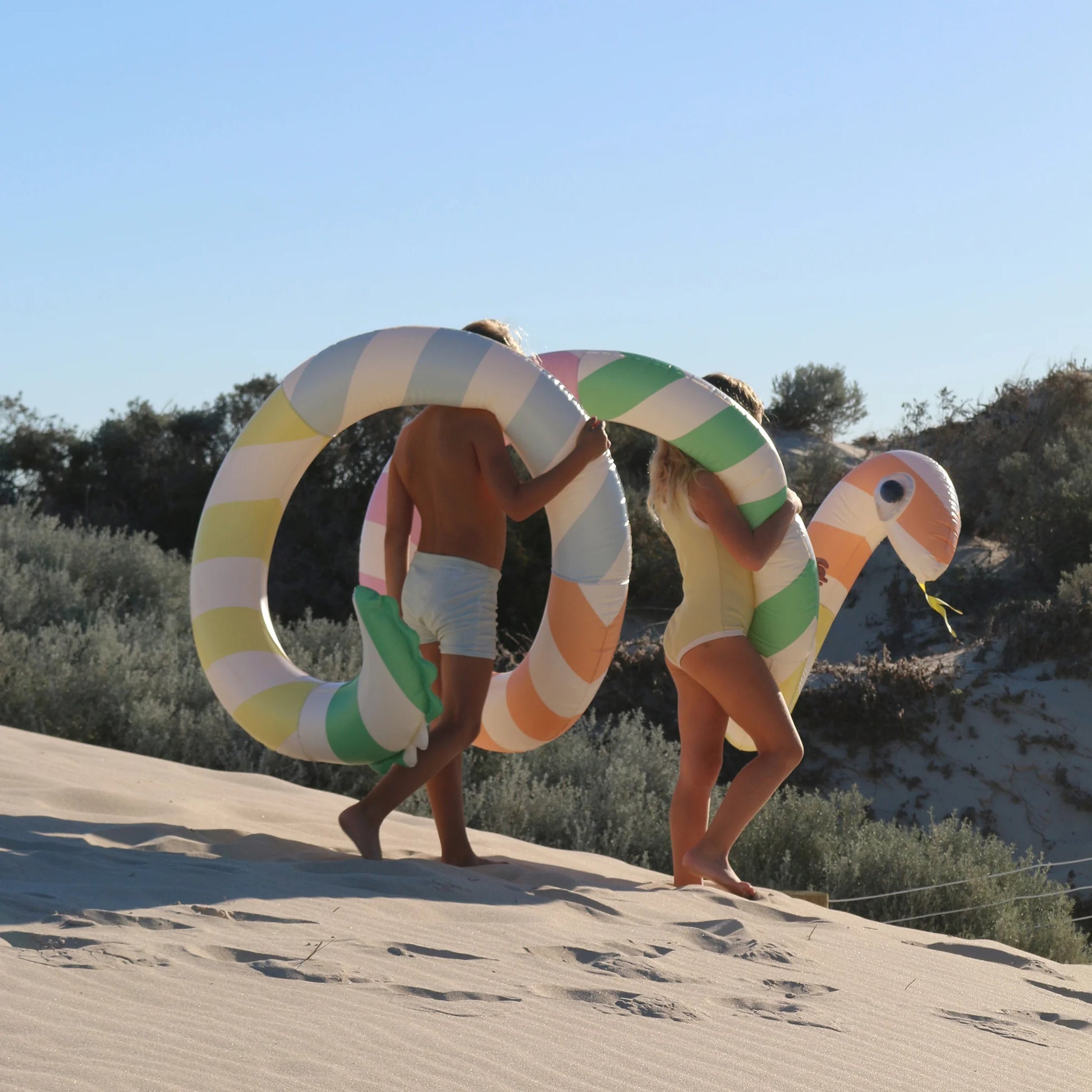 Gigantyczny dmuchany makaron w kolorowe paski niesiony przez dwoje ludzi na plaży LOQI