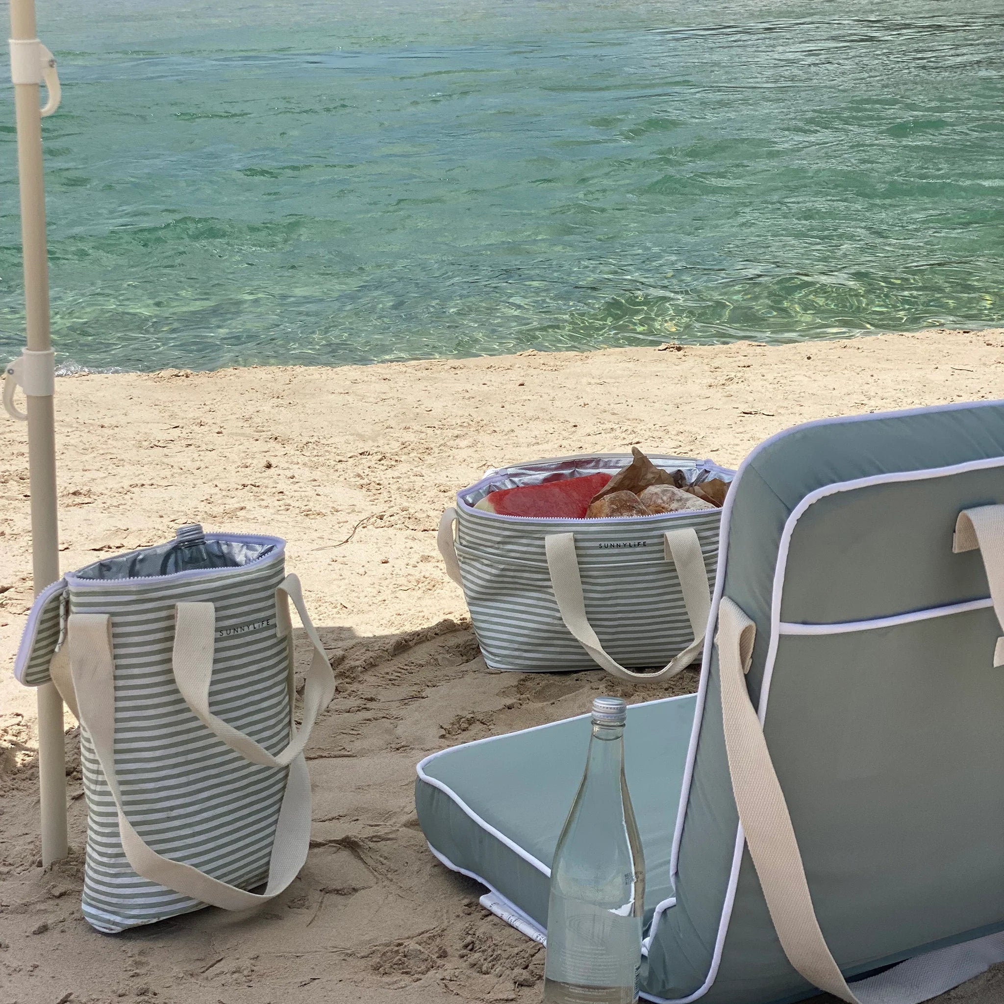 Torba termiczna na lunch LOQI w paski na plaży obok turkusowego krzesła i butelki wody
