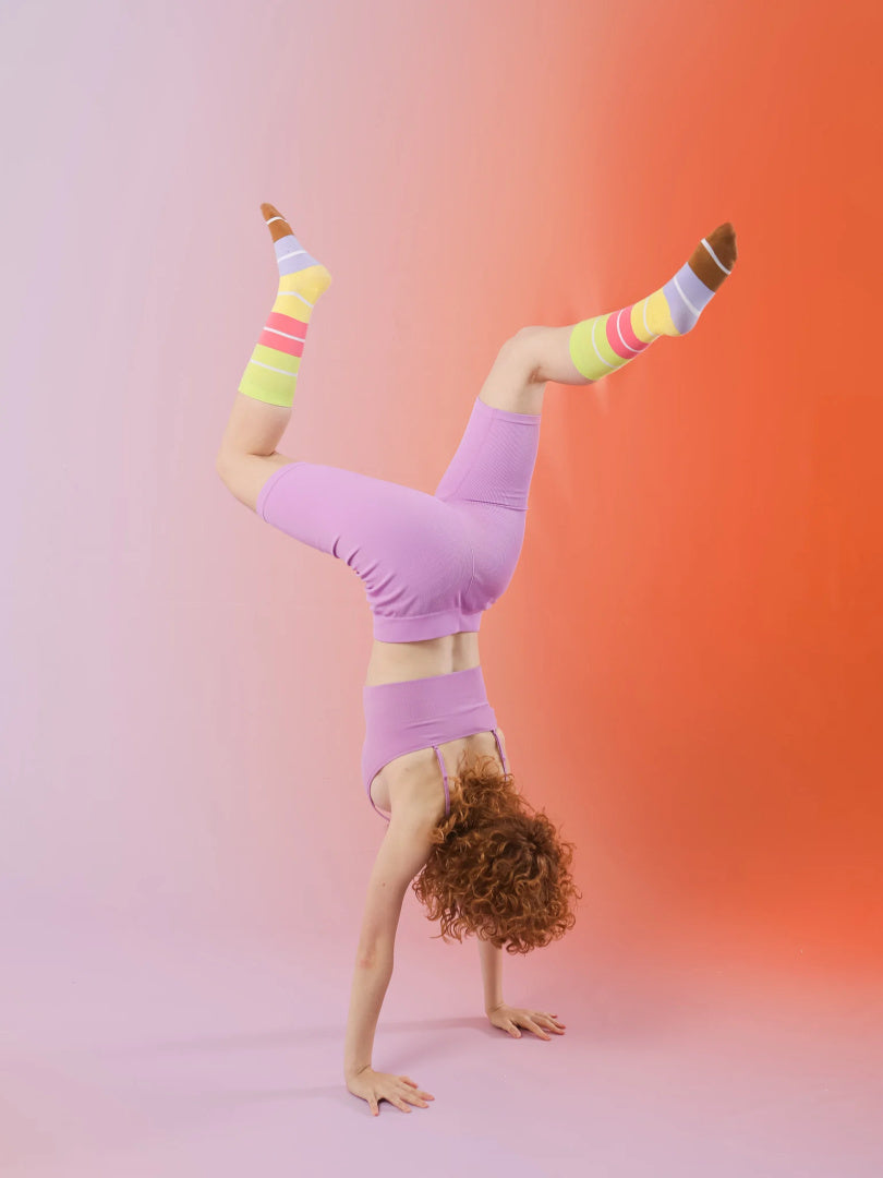 Kobieta w kolorowych skarpetkach Bon Macaron wykonuje stojak na rękach na gradientowym tle