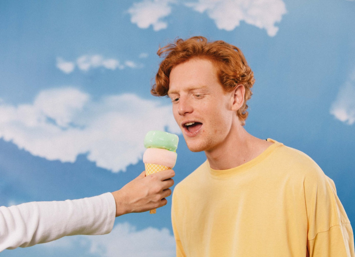 Mężczyzna w żółtej koszulce bawi się z ręką trzymającą kolorowe skarpetki w kształcie lodów na tle nieba