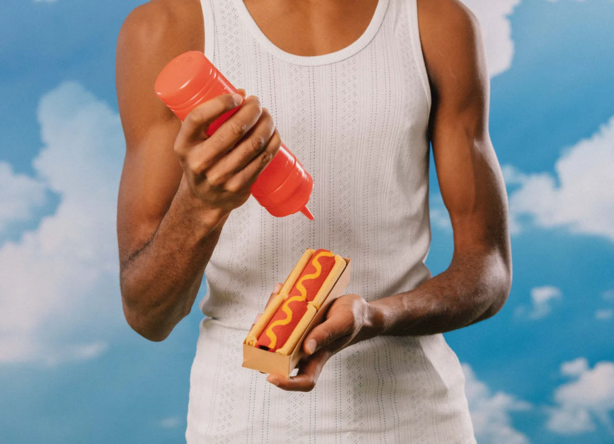 Mężczyzna w białej koszulce bez rękawów trzymający hot doga z ketchupem na tle nieba