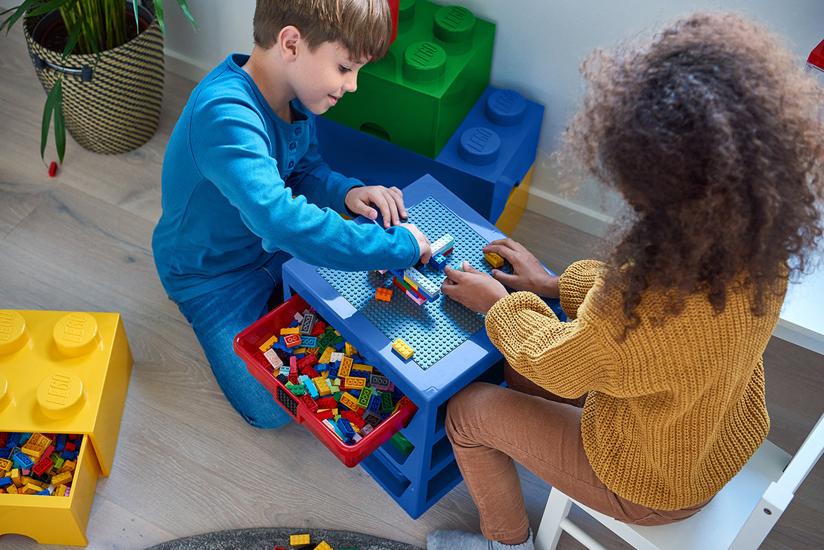 Dwoje dzieci bawi się przy stole z niebieską szufladą klocek LEGO pełną kolorowych elementów