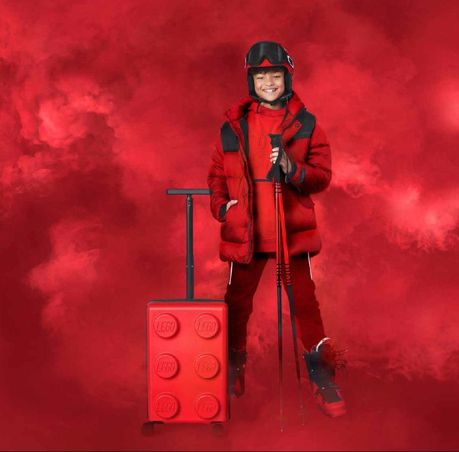 Dziecko w czerwonym ubraniu narciarskim z walizką LEGO Signature czerwona na tle czerwonego dymu