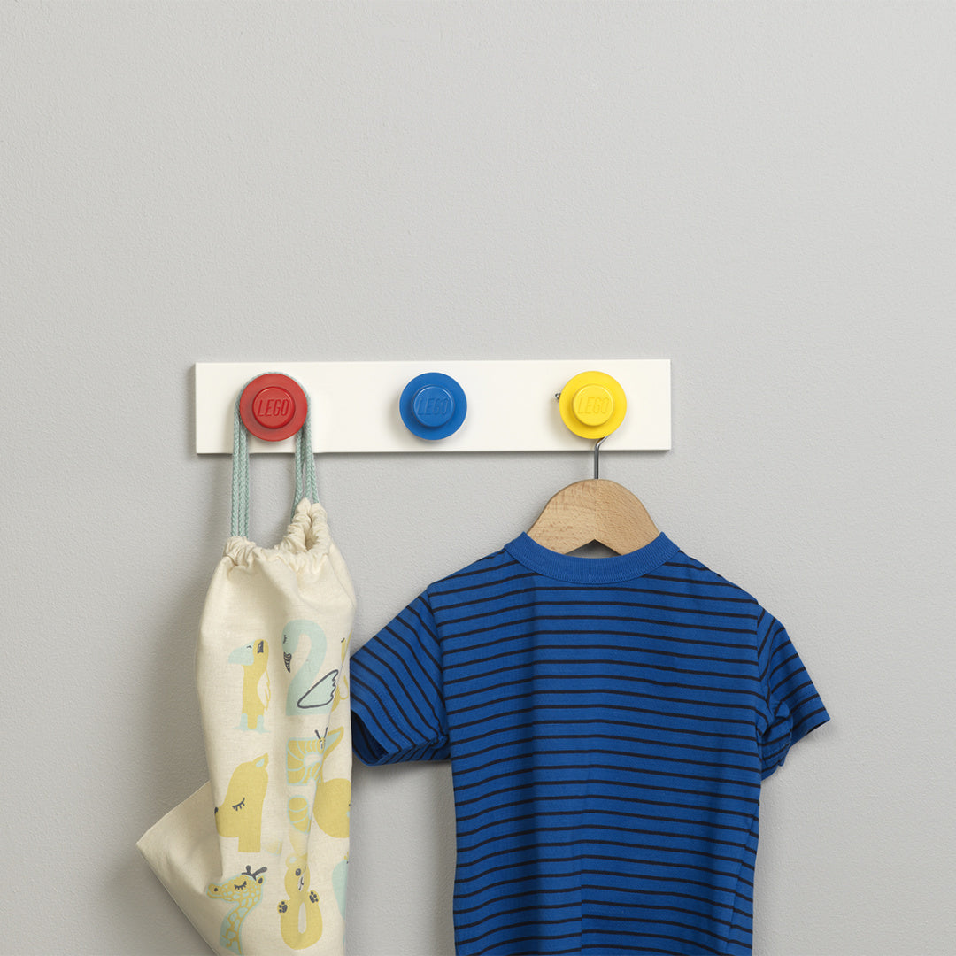 Wieszaki LEGO na ścianę w kolorach czerwonym, niebieskim i żółtym z ubraniami i torebką zawieszonymi na białej desce