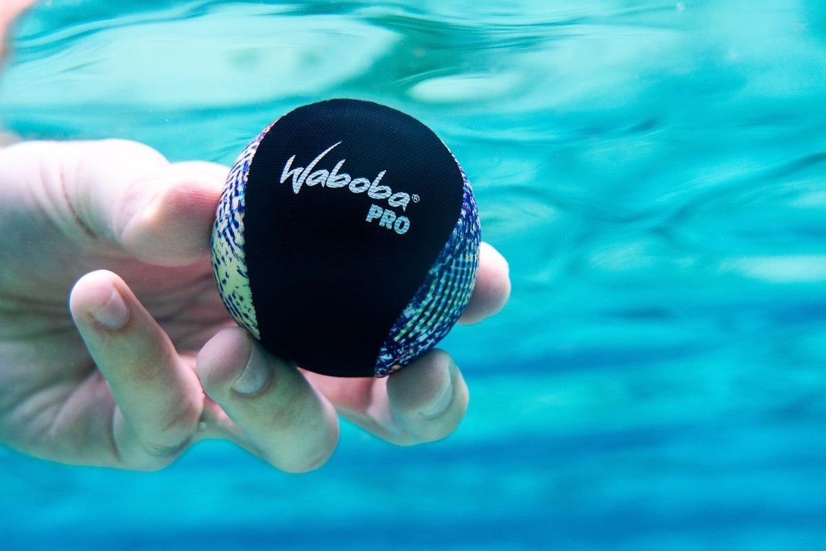 Piłeczka Waboba Pro do odbijania na wodzie trzymana ręką pod wodą na niebieskim tle