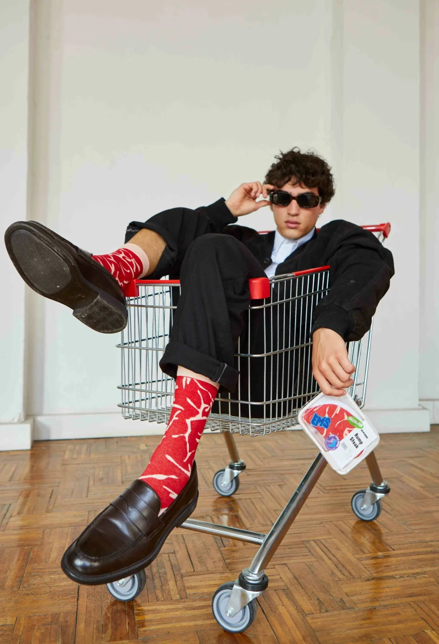 Młody mężczyzna w czarnych okularach i skarpetkach steak dla dorosłych siedzi w wózku na zakupy