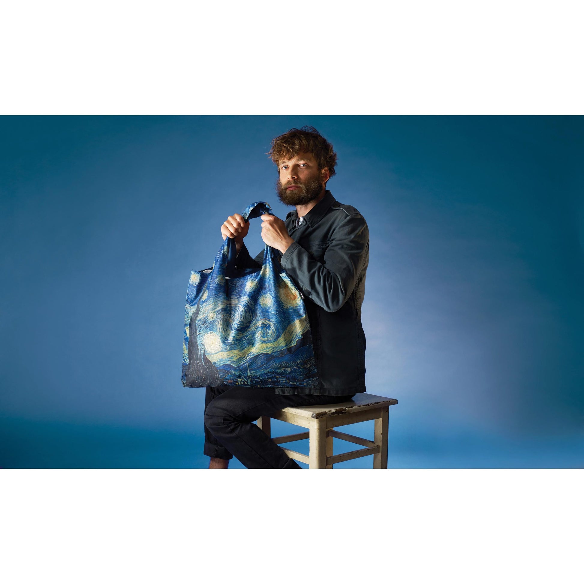 Mężczyzna siedzi na krześle z torbą podręczną Vincent van Gogh przedstawiającą obraz Gwiaździsta noc