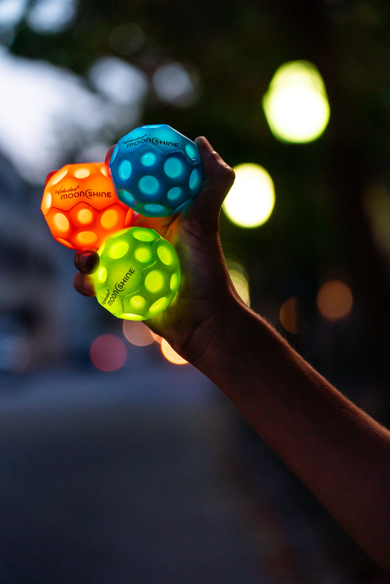 Ręka trzymająca piłeczkę Waboba Moonshine niebieska wraz z pomarańczową i zieloną piłeczką na rozmytym tle