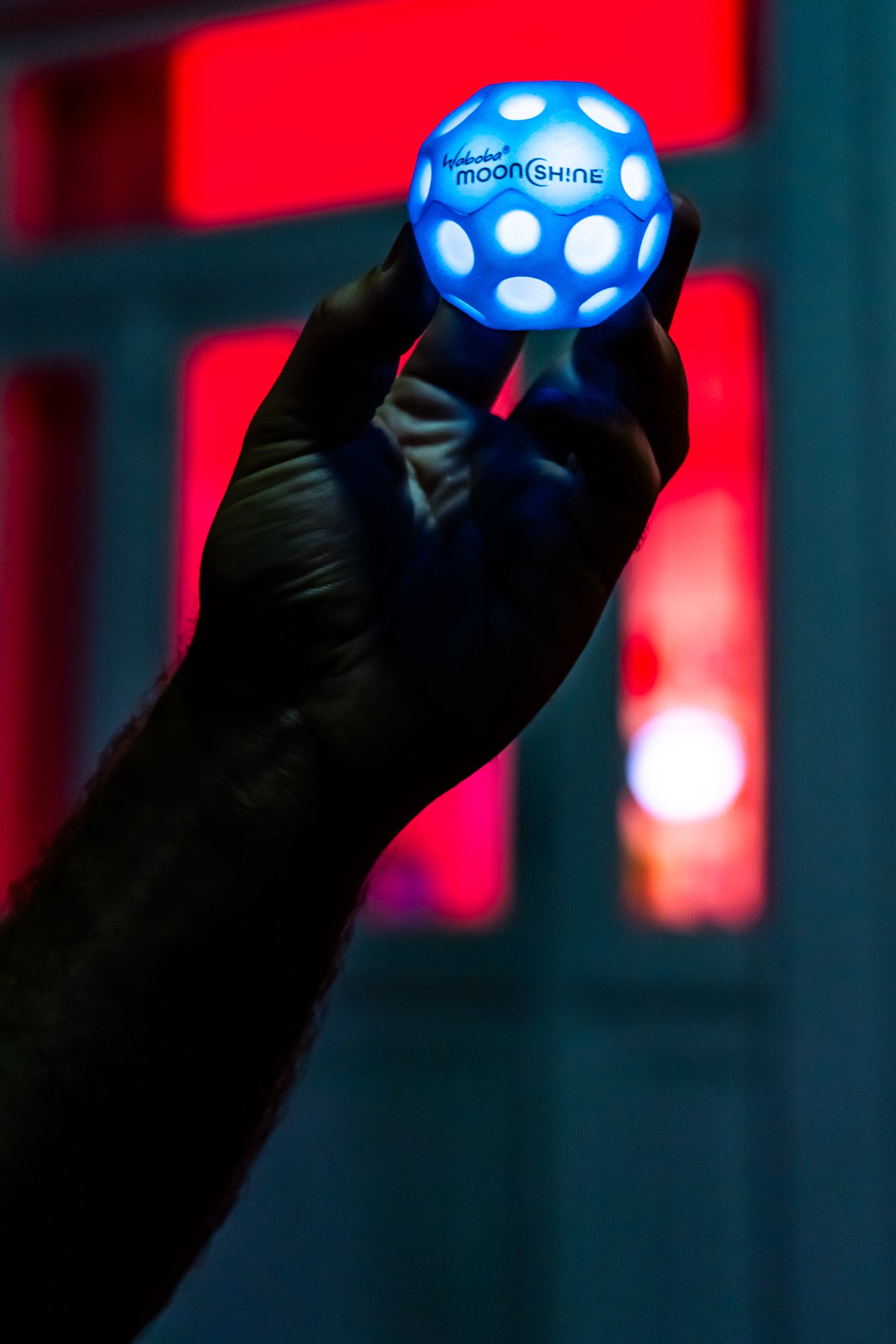 Piłeczka Waboba Moonshine niebieska świecąca w ciemności trzymana w ręce na tle czerwonego i niebieskiego światła