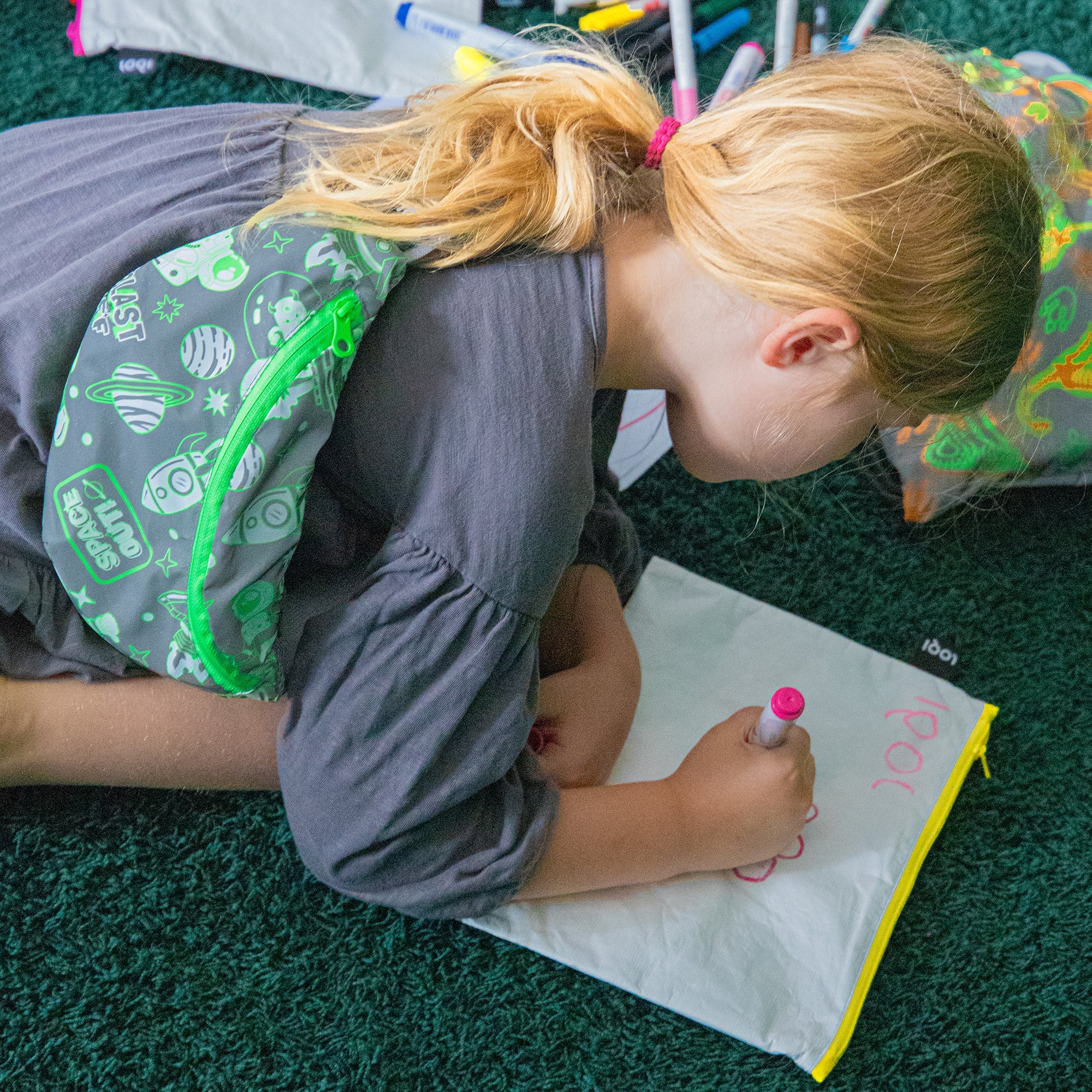 Dziewczynka z blond włosami leżąca na dywanie z zieloną nerką odblaskową marki Yamann, pisząca na białej tablicy