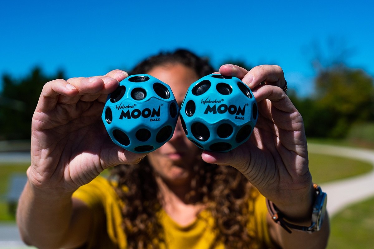 Dwie niebieskie piłeczki Waboba Moon Ball trzymane w dłoniach na tle zielonego parku