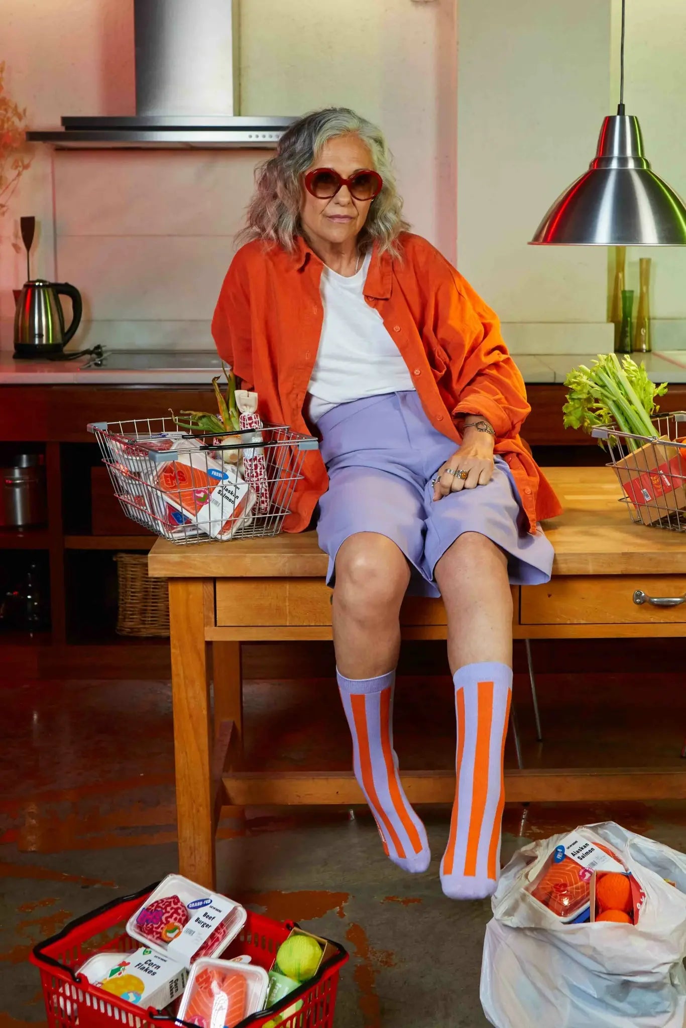 Skarpetki łosoś Eat My Socks na starszej kobiecie siedzącej na stole w kuchni ubranej w pomarańczową koszulę i fioletowe spodenki
