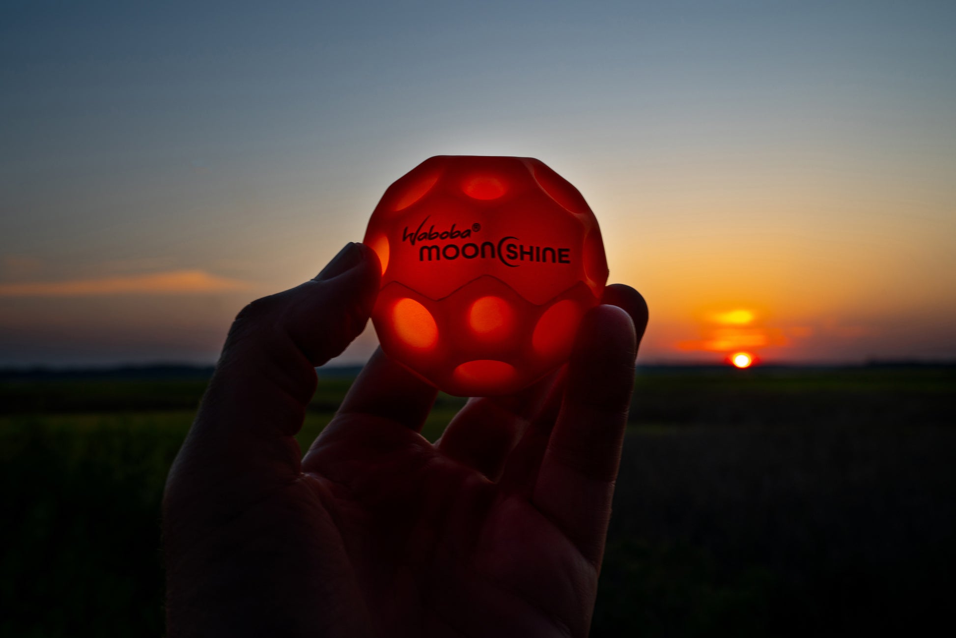 Czerwona piłeczka świecąca Waboba Moonshine trzymana na tle zachodu słońca na zewnątrz
