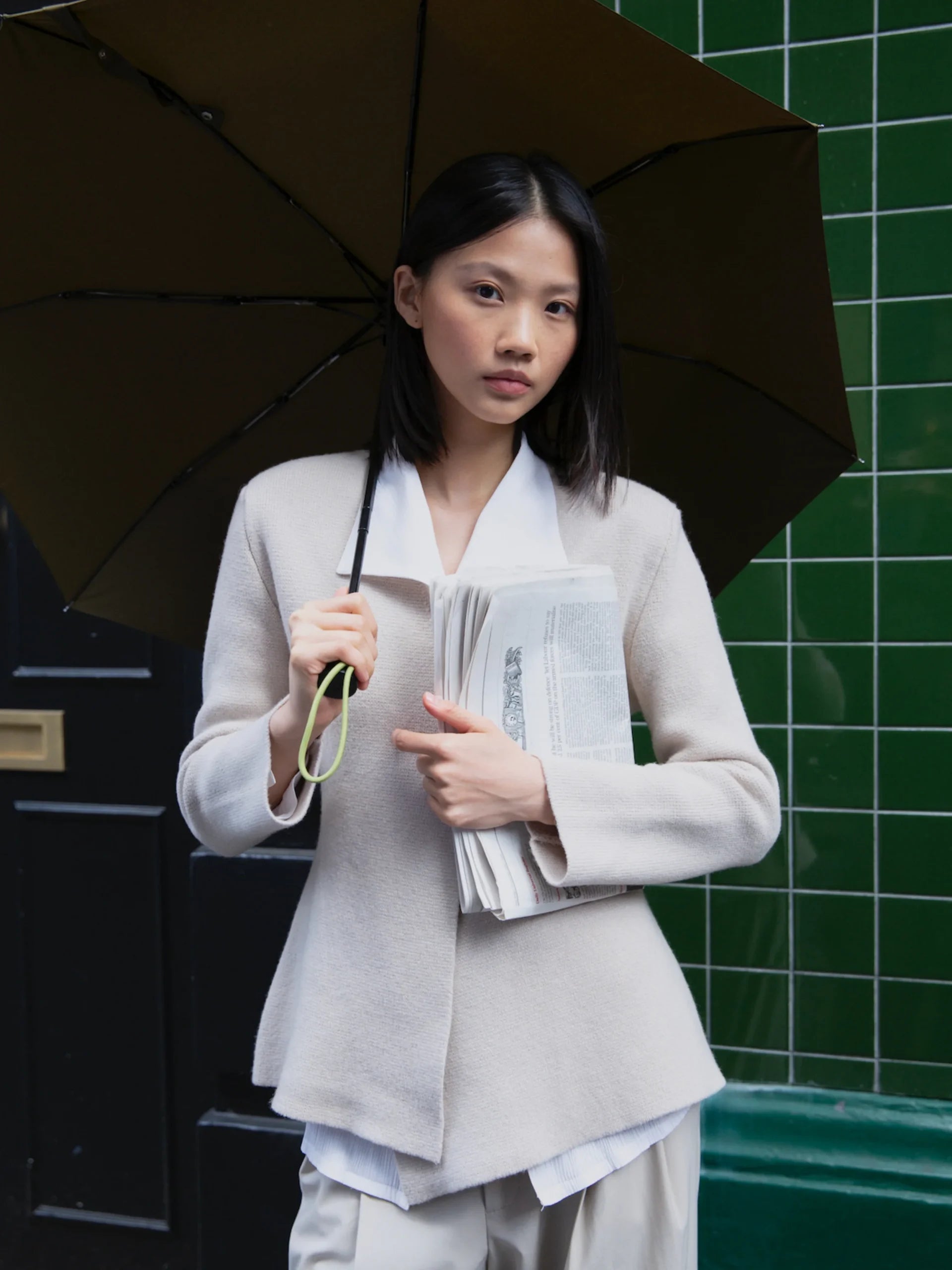 Kobieta trzymająca parasolkę automatyczną ciemną oliwkową oraz gazetę na tle zielonych płytek