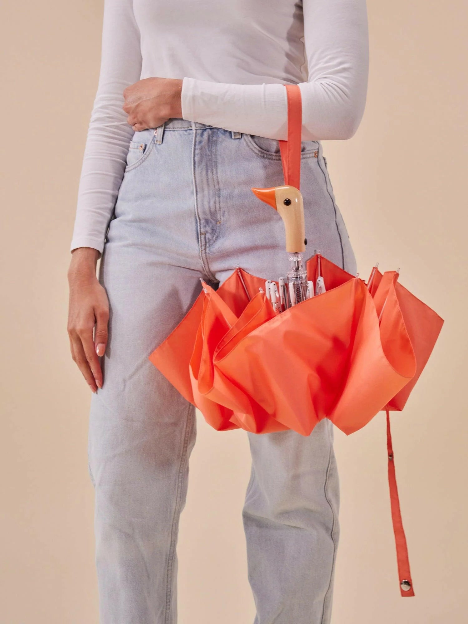 Pomarańczowa parasolka z rączką w kształcie kaczego dzioba trzymana przez osobę w jasnych spodniach i białej bluzce