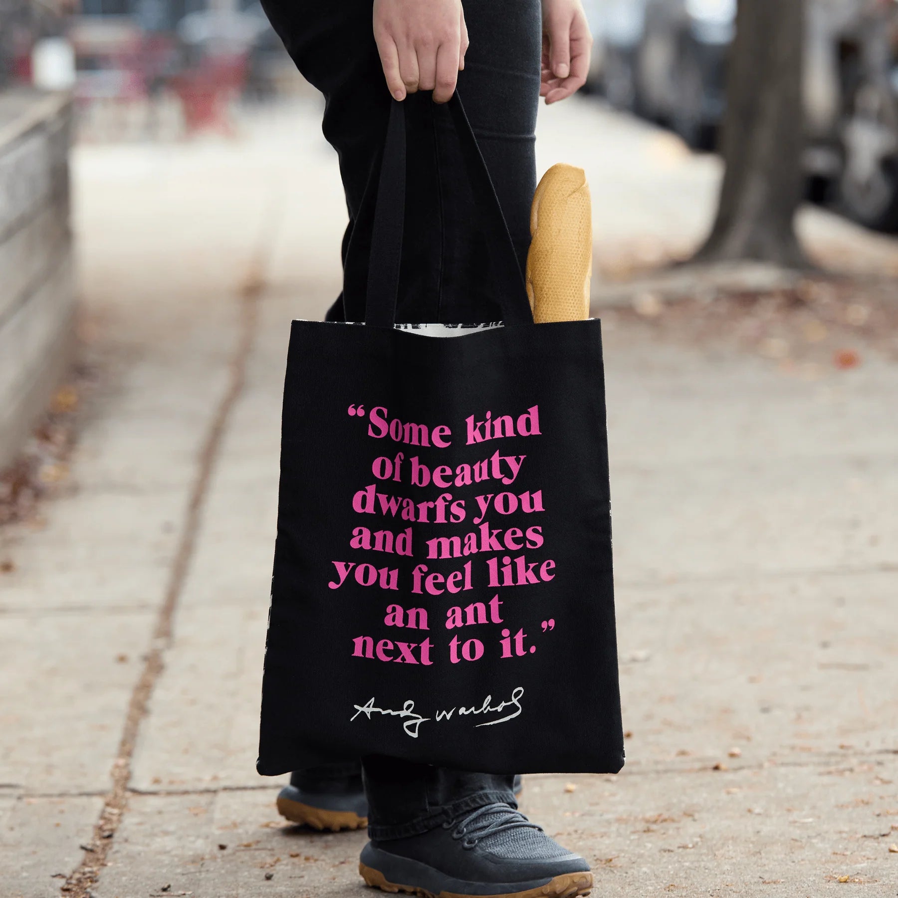 Czarna torba bawełniana Andy Warhol z różowym napisem trzymana na chodniku z bagietką w środku