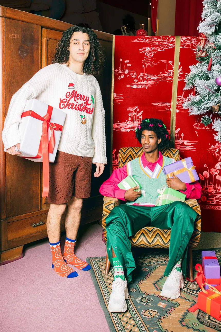 Mężczyzna w skarpetkach świąteczne ciasteczko stoi obok drugiego mężczyzny trzymającego prezenty w świątecznym wnętrzu