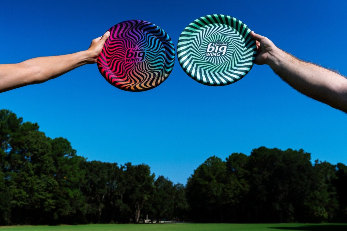 Dwa kolorowe latające dyski Waboba Bigwing trzymane przez dwie osoby na tle niebieskiego nieba i zielonych drzew