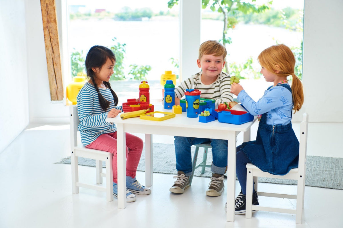 Trójka dzieci siedzi przy białym stoliku z akcesoriami LEGO i bidon LEGO dla chłopca