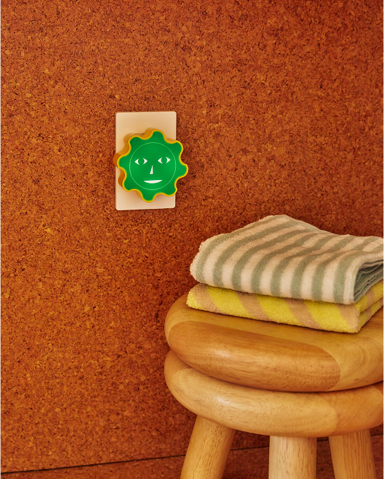 Lampka nocna dla dzieci Areaware w kształcie zielonego uśmiechniętego słońca podłączona do gniazdka na tle korkowej ściany