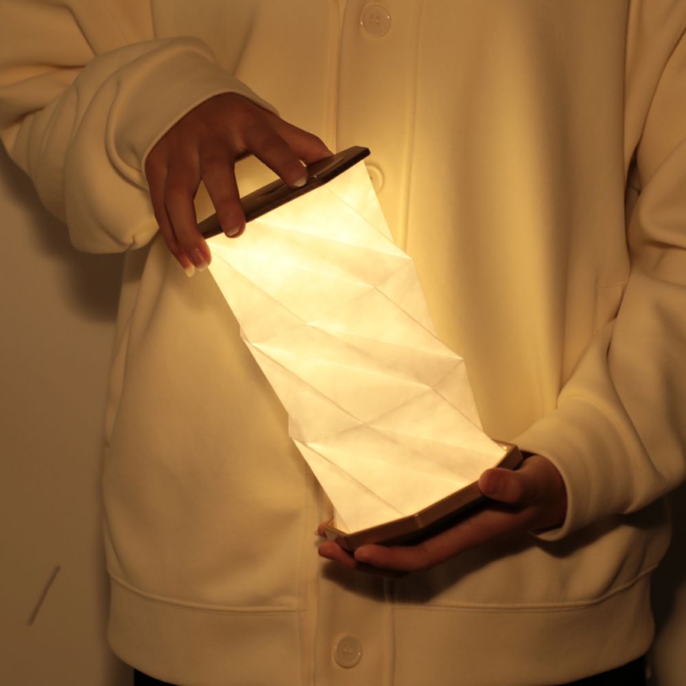 Osoba trzymająca świecącą sześciokątną lampę stołową Gingko Design o unikalnym składanym kształcie