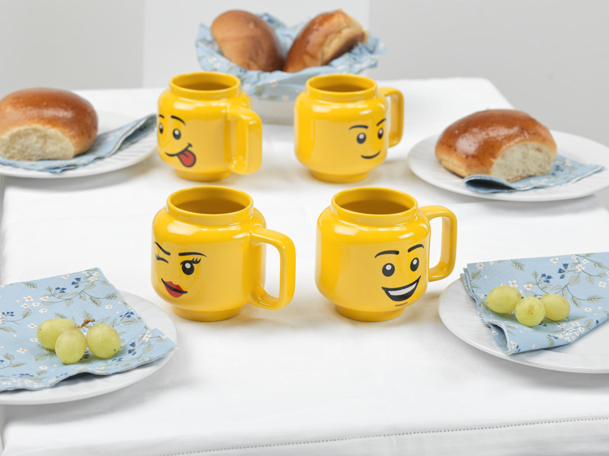 kubek LEGO ceramiczny w żółtym kolorze z różnymi wyrazami twarzy na stole z bułkami i winogronami