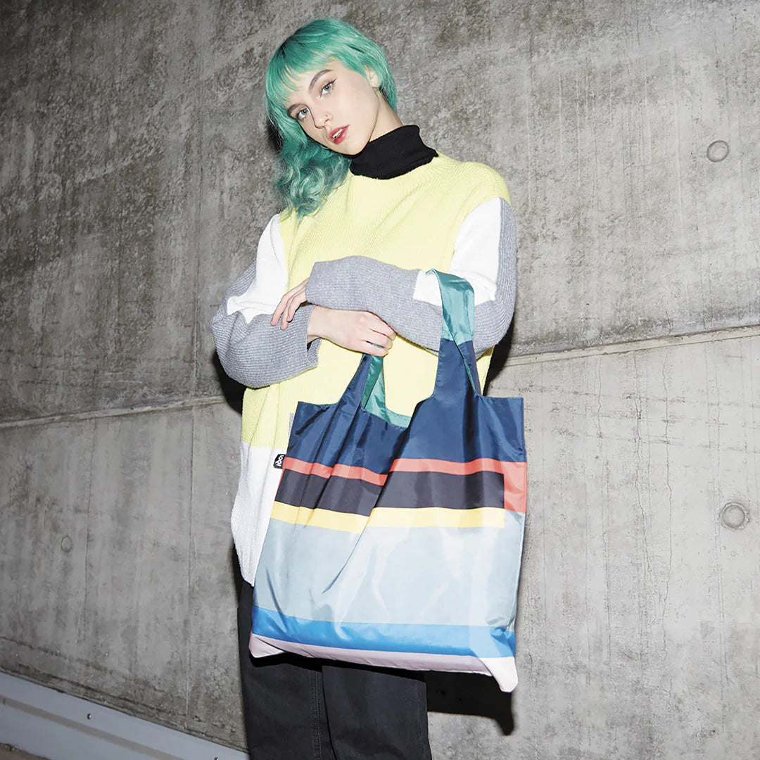 Torba LOQI Jean Spencer z kolorowymi poziomymi paskami trzymana przez osobę na tle betonowej ściany